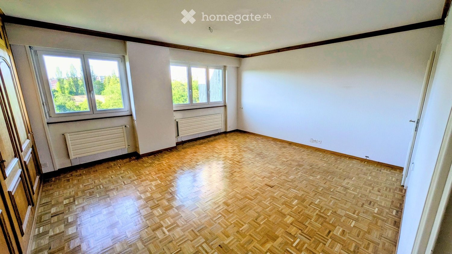 Empty room with wooden parquet flooring, white walls, and large windows offering a view of greenery outside. Radiators are visible beneath the windows.