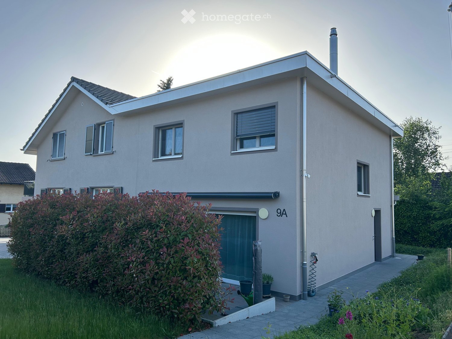 Two-story house with light gray exterior, multiple windows, and a small garden area with potted plants and shrubs.