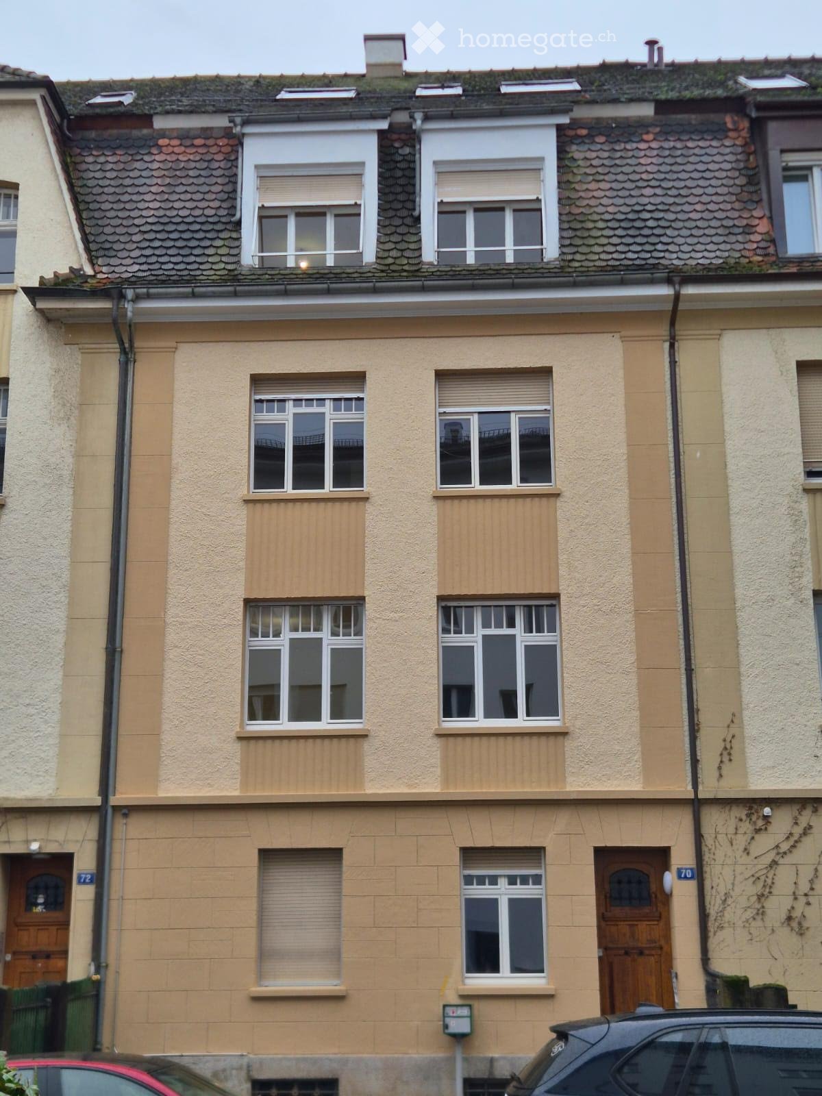 Two-story residential building with beige facade, multiple windows with shutters, and wooden doors.