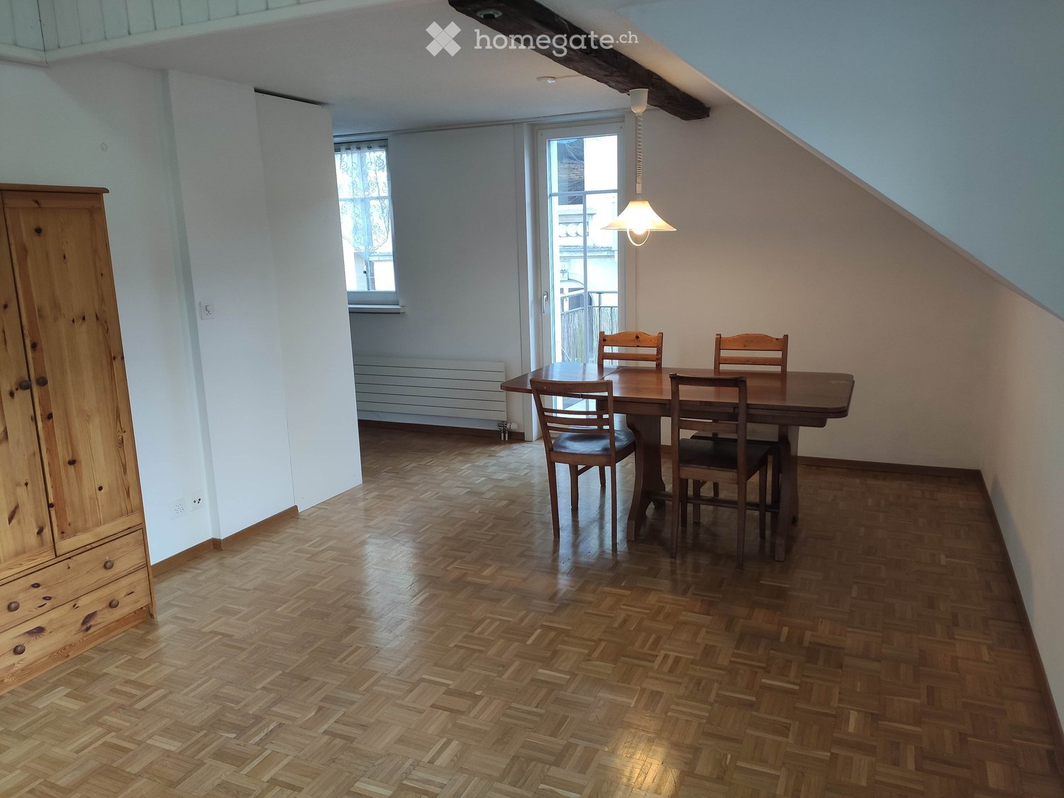Bright room with wooden herringbone flooring, a large wooden table with four chairs, and a tall wooden wardrobe. The room has white walls, a slanted ceiling, and a hanging light fixture.