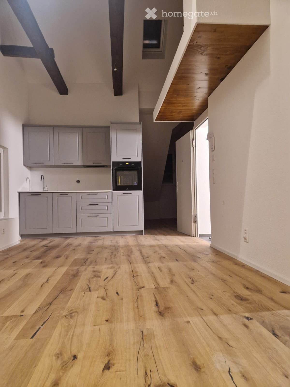 Empty kitchen with light grey cabinets, stainless steel sink, and built-in oven. The floor is made of light wood with visible grain patterns. The space is open and well-lit with natural light coming from a window on the left side.