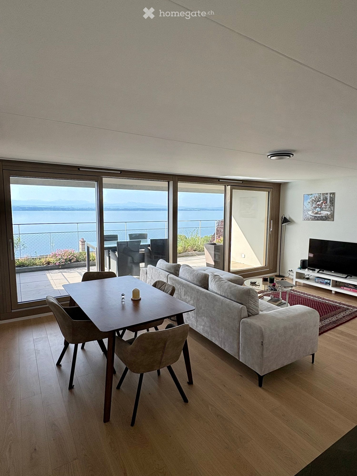 Modern living room with large windows offering a scenic view, featuring a grey sofa, wooden dining table, chairs, and a television on a stand.