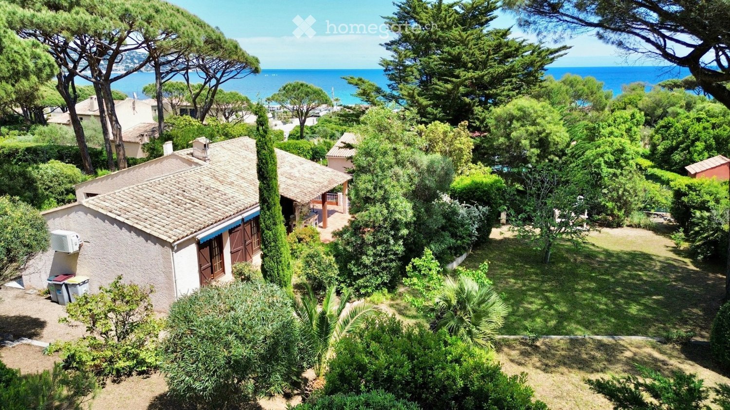 Aerial view of a coastal garden area with a small house, surrounded by lush greenery and trees, overlooking the sea.