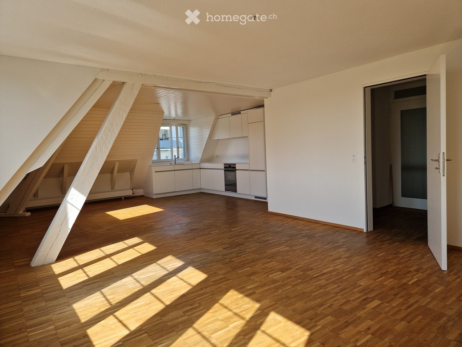 Bright, empty attic room with wooden flooring, white walls, and exposed wooden beams. Sunlight streams in through a window, casting shadows on the floor. A doorway leads to a small, white kitchen area with cabinets and a sink.