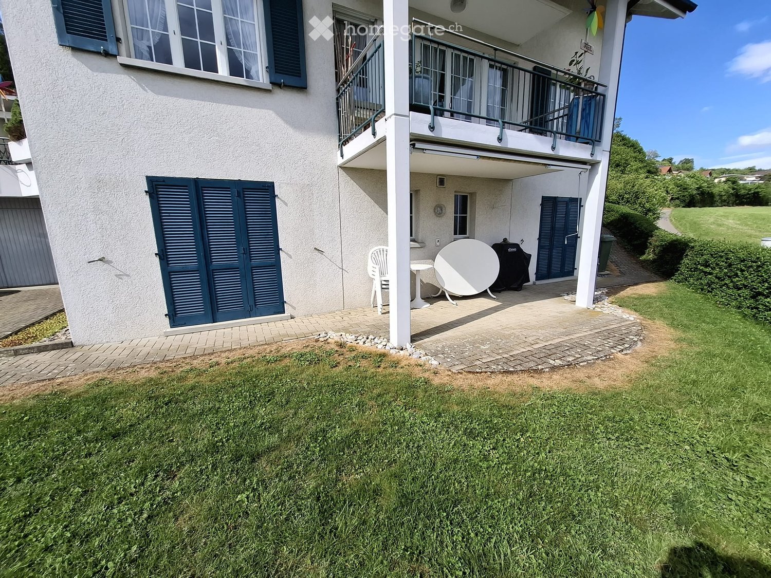 A well-maintained garden with a paved patio area, featuring a white table, a chair, and a satellite dish. The house has multiple windows with shutters and a balcony with railings.