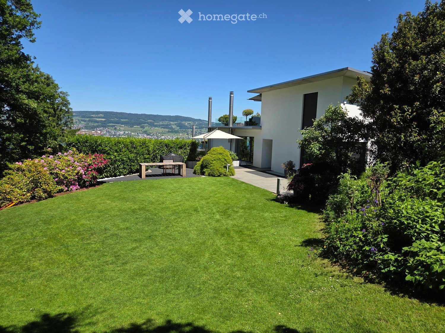 Well-maintained lawn with surrounding greenery, a modern white house with large windows, a patio area with a table and chairs, and a scenic view of the countryside.