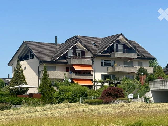 Two-story house with a dark tiled roof, multiple balconies, and orange awnings. The house is surrounded by trees and a well-maintained garden.