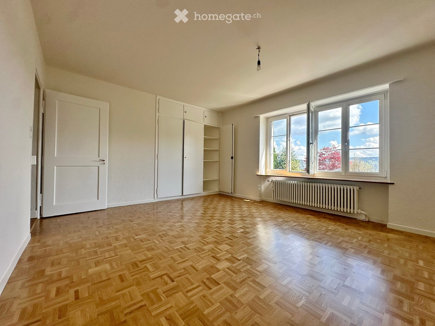 Empty room with wooden parquet flooring, white walls, large windows, white built-in wardrobes, white radiator, and a ceiling light.