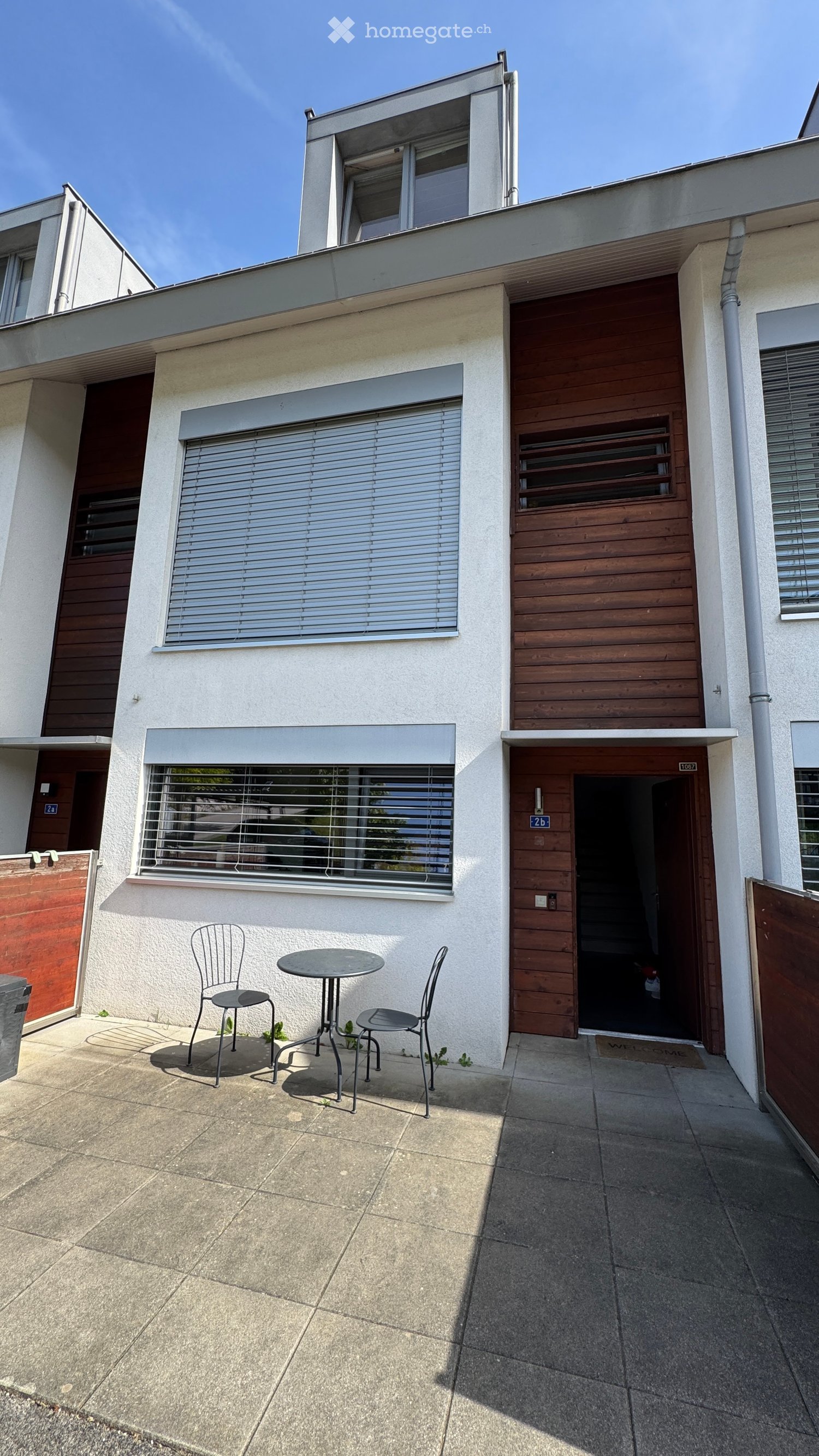 Small outdoor seating area with a round table and two chairs, adjacent to a two-story building with a garage and covered entrance.