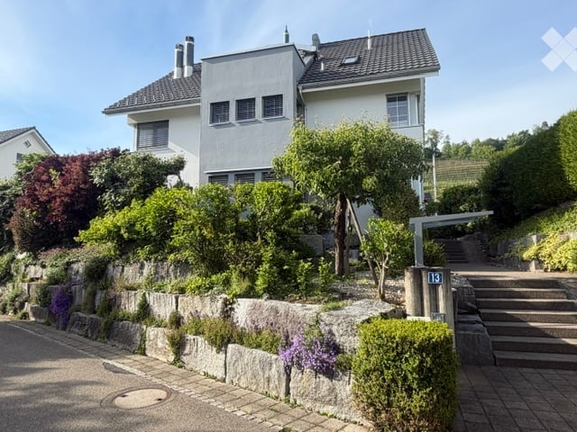 Two-story house with a modern design, surrounded by greenery and a stone retaining wall. The house features a flat roof, multiple windows, and a small staircase leading to the entrance.