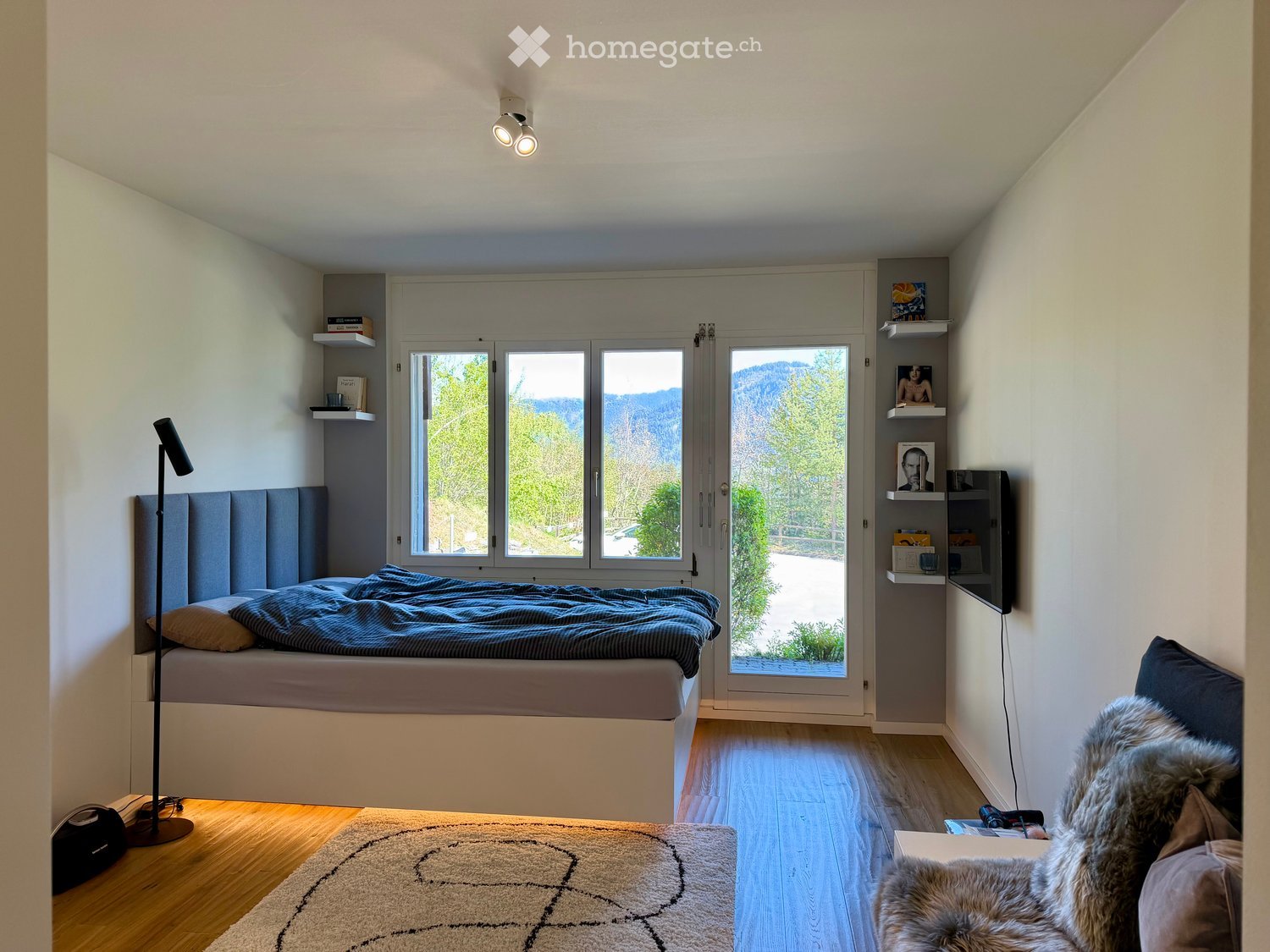 Modern bedroom with a large window offering a scenic mountain view, featuring a bed with a headboard, a floor lamp, shelves, a fur chair, and a patterned rug.