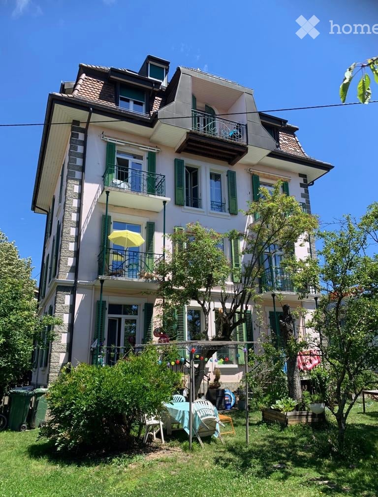 A multi-story house with green shutters, balconies, and a well-maintained garden with outdoor furniture.