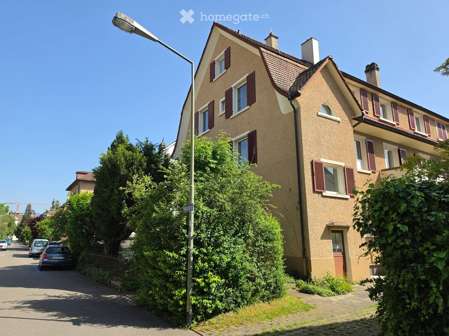 Two-story house with beige siding, red shutters, multiple windows, and a chimney. Surrounded by greenery and located on a quiet street.
