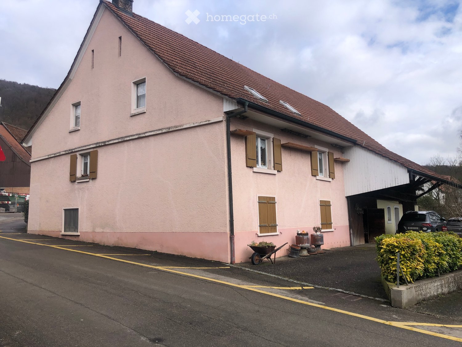 Two-story house with pink exterior, multiple windows with shutters, garage, and small garden area with potted plants and a wheelbarrow.