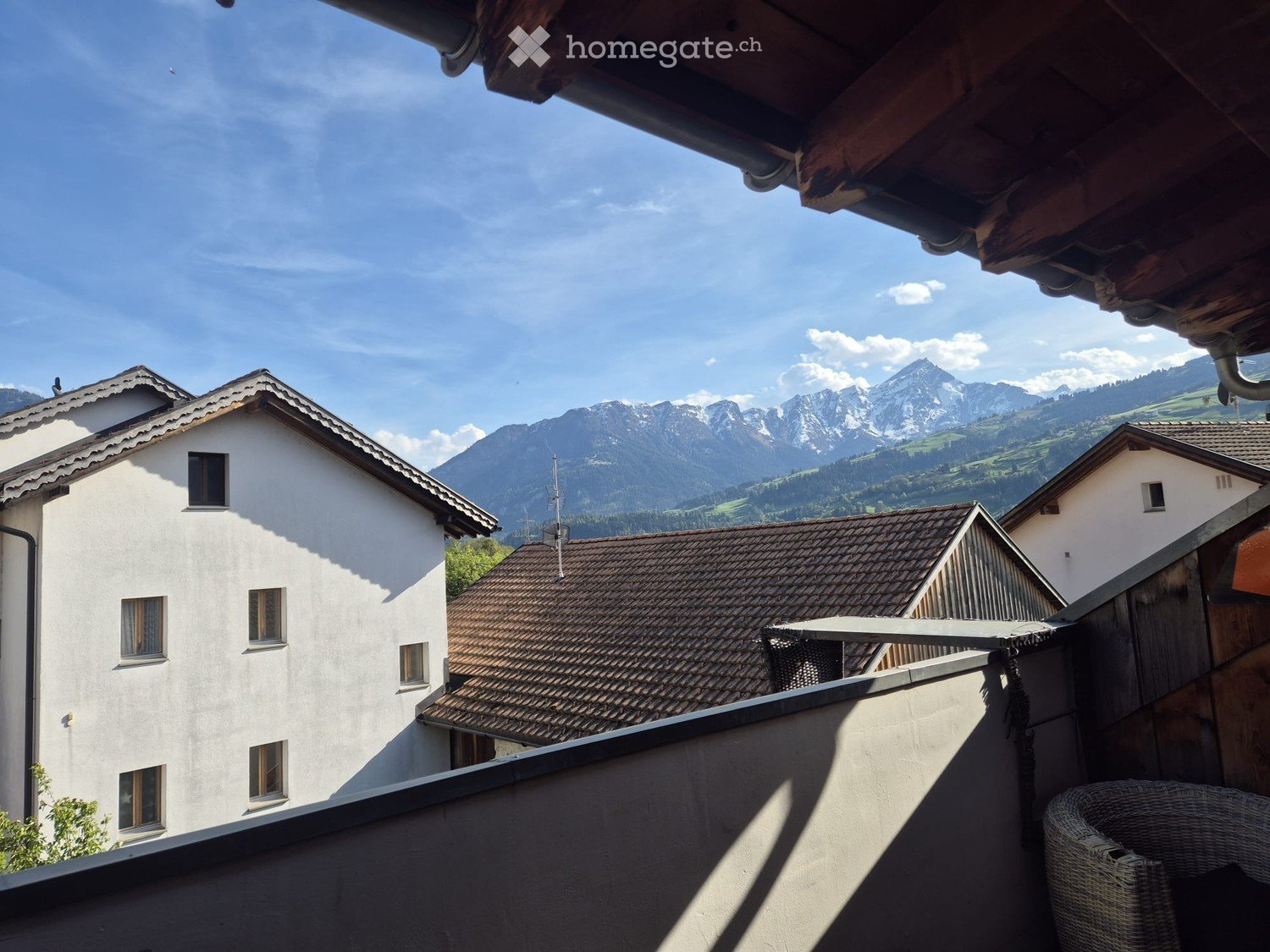 Balcony with a wicker chair, overlooking a village with white houses and distant mountain ranges under a clear blue sky.