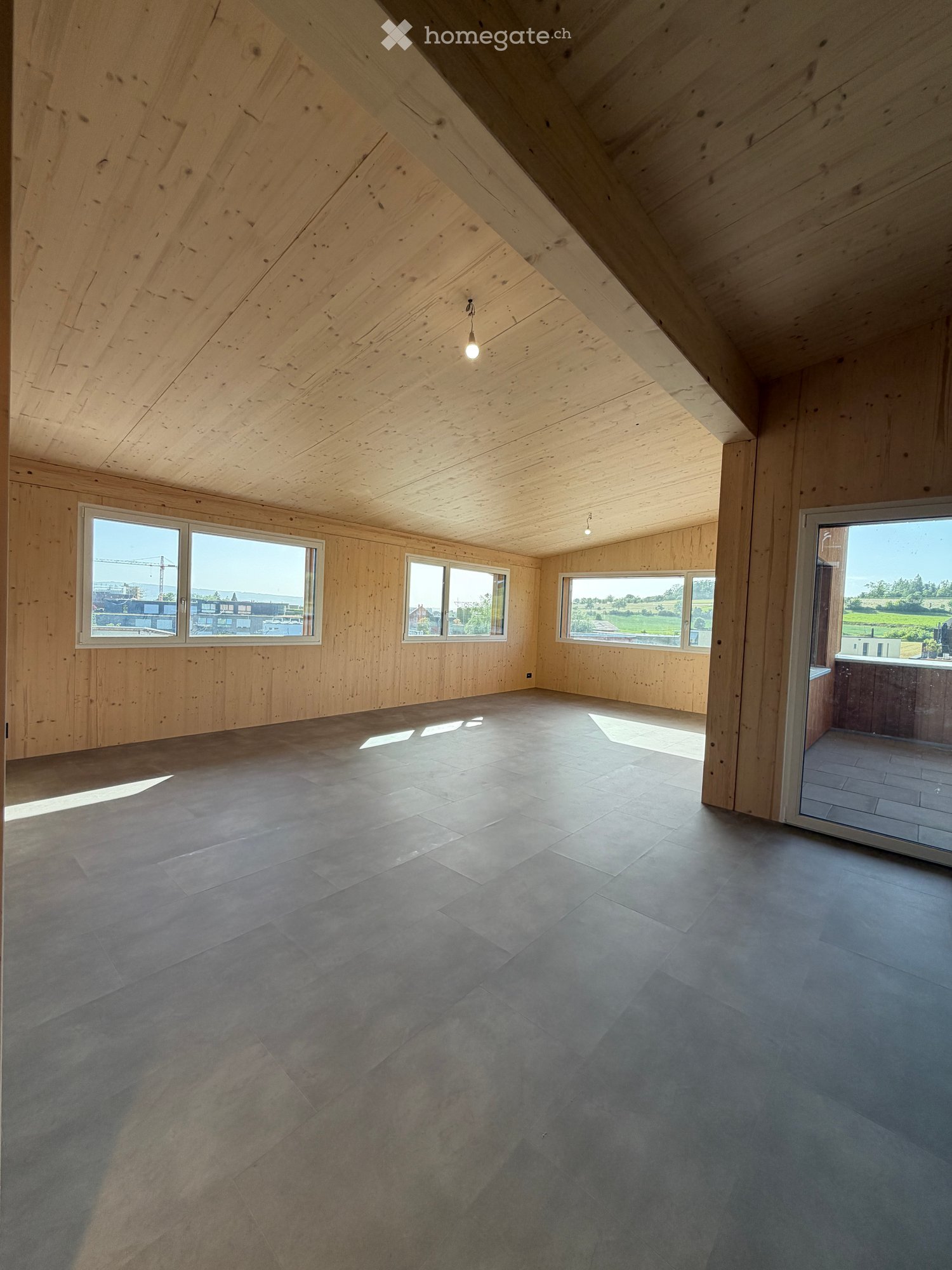 An empty attic space with wooden walls and ceiling, large windows, and a sliding glass door leading to a balcony.