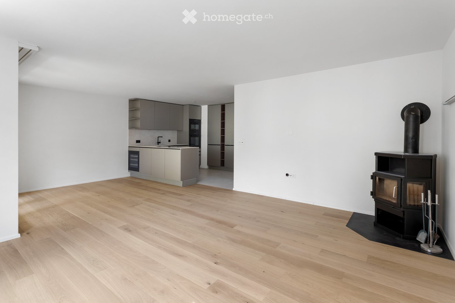 Open-plan living and kitchen area with light wooden flooring, modern grey kitchen cabinets, a central island with sink and faucet, built-in oven, refrigerator, and a wood-burning stove with a chimney.