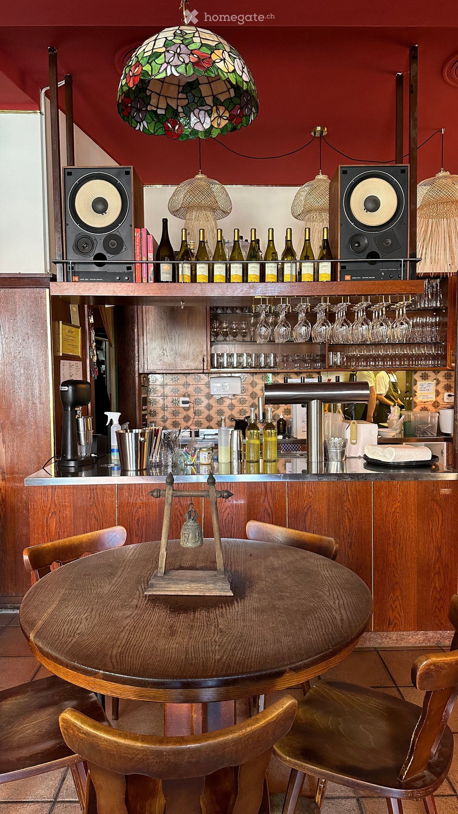 A wooden table with chairs, a wooden bar counter, a shelf with bottles, wine glasses, and a lamp