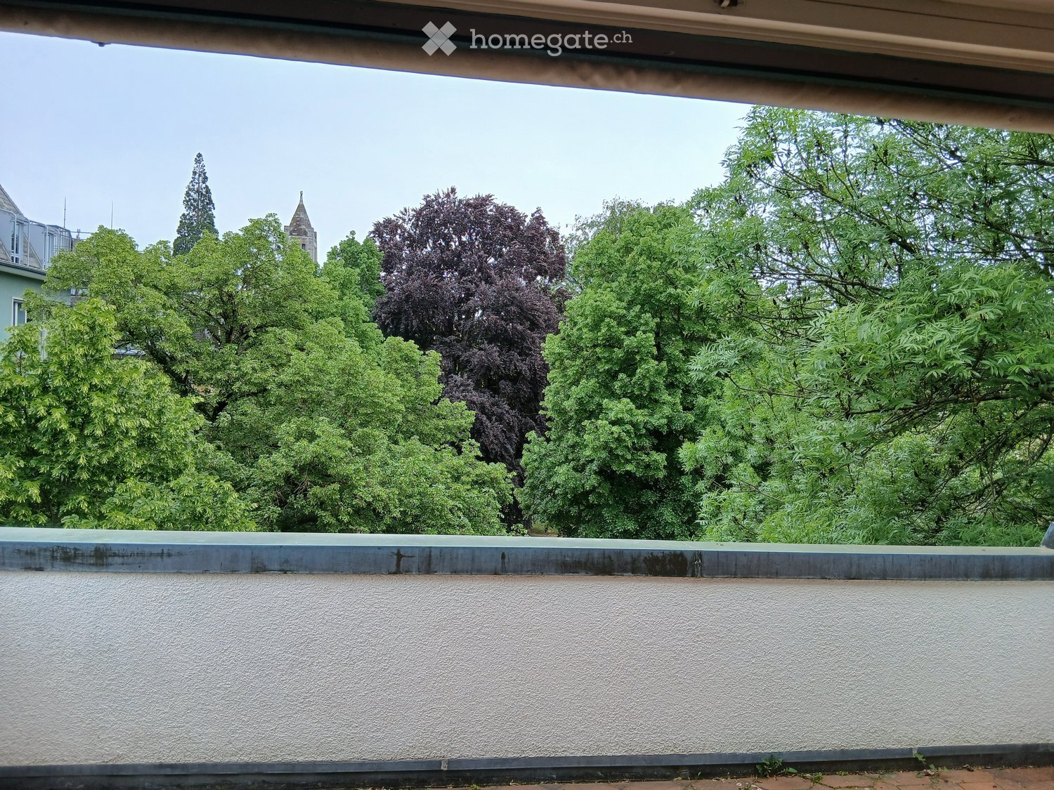 Balcony with a low concrete wall, overlooking a lush green area with trees and a distant building with a spire.