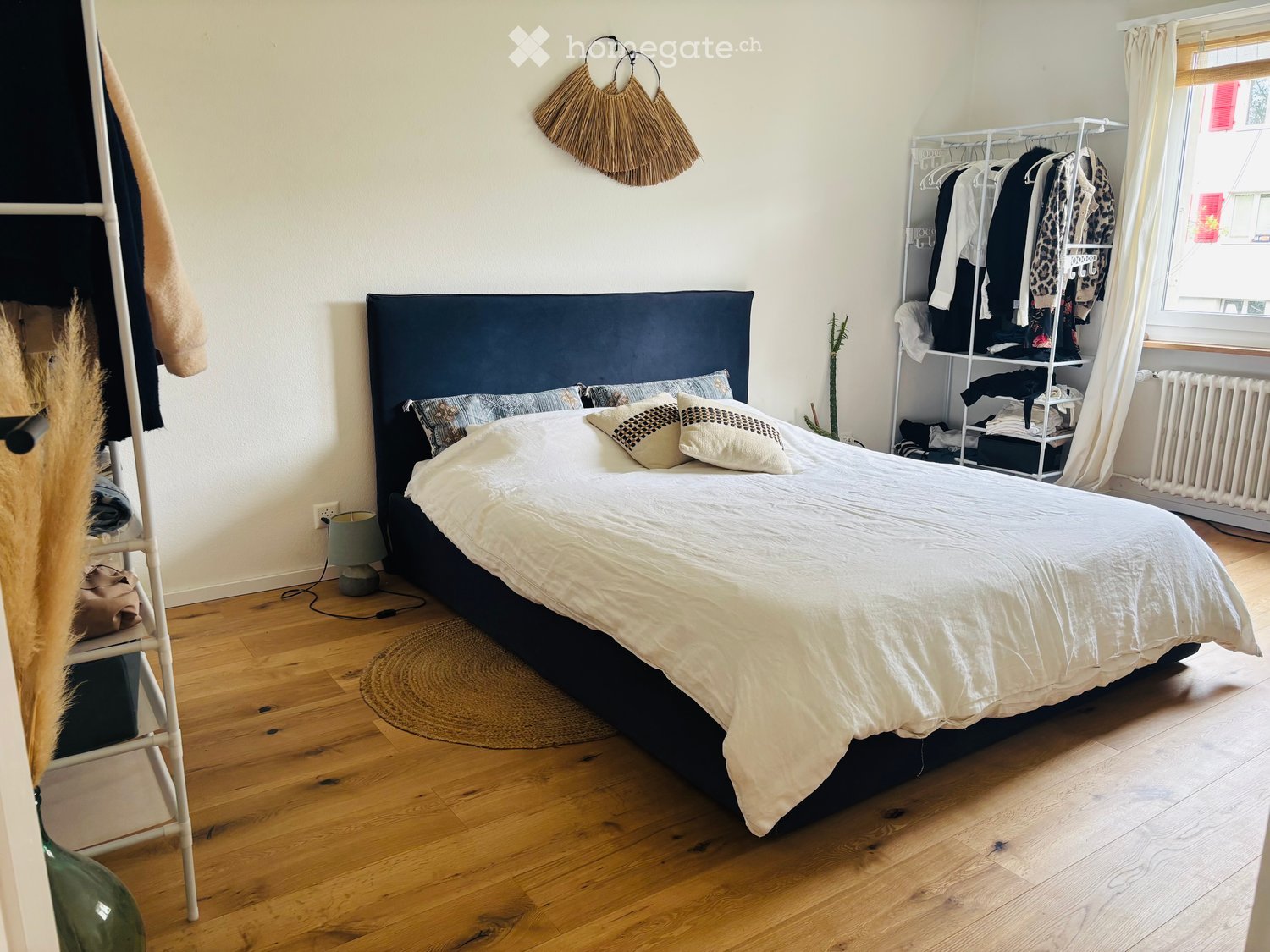 Cozy bedroom with wooden flooring, a large bed with white bedding and decorative pillows, a bedside lamp, a white clothing rack, and a woven wall hanging.