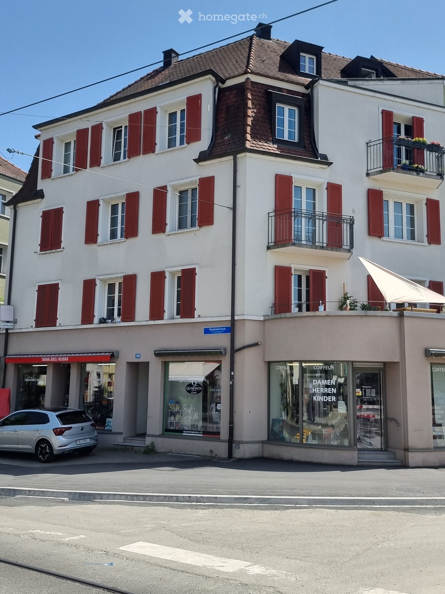 Three-story building with white facade, red shutters, multiple windows, and commercial storefronts on the ground floor. Includes a balcony on the second floor and a small awning over one of the entrances.
