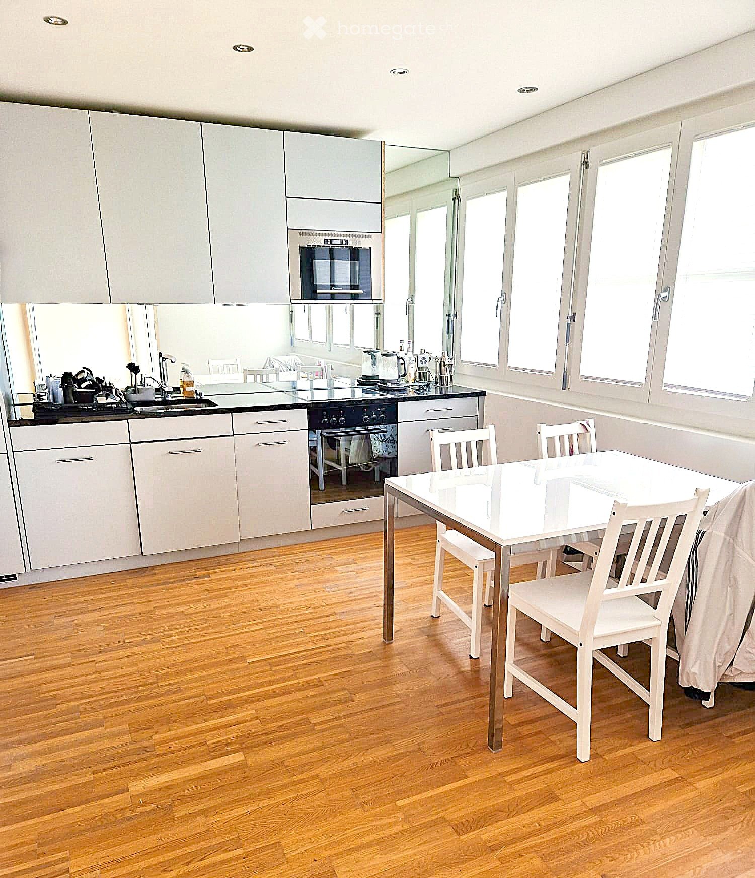 Modern kitchen with light-colored cabinets, a white dining table with four chairs, and various appliances including an oven, stove, and coffee maker. The room features wooden flooring and large windows.