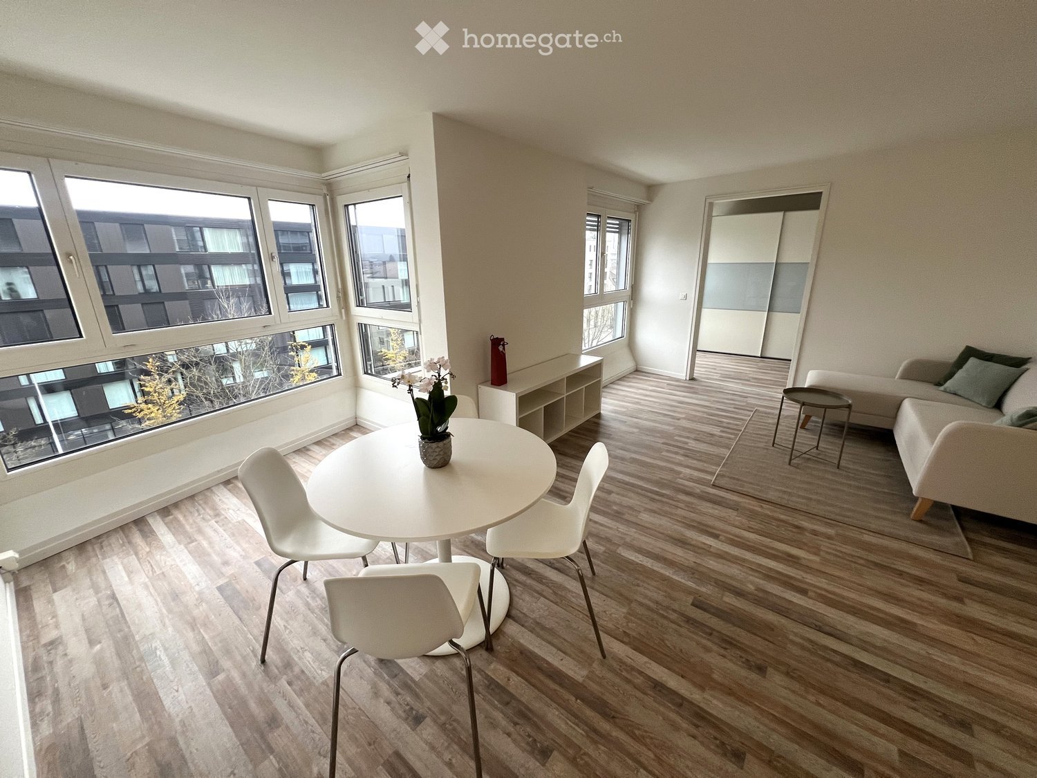 Modern living room with light wooden flooring, large windows, a white dining table with chairs, a sofa, and a small shelf with a flower pot.