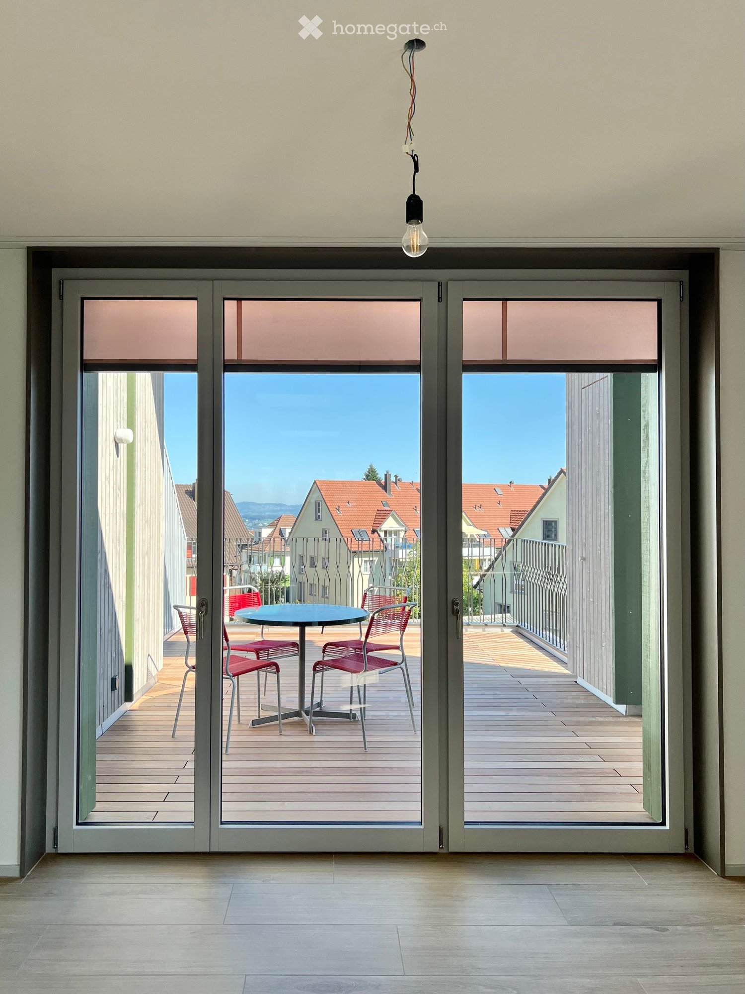 Balcony with wooden decking, table, and chairs, overlooking a residential area with red-roofed houses and a clear blue sky.