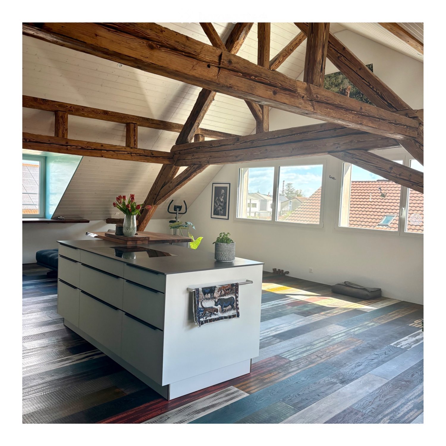 Modern kitchen with exposed wooden beams, white cabinets, island with sink, and colorful patterned rug.