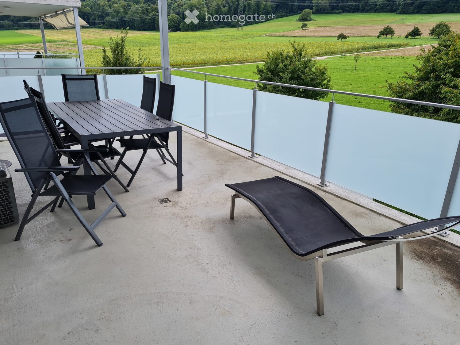 Balcony with a table, chairs, and a lounge chair, overlooking a green field.