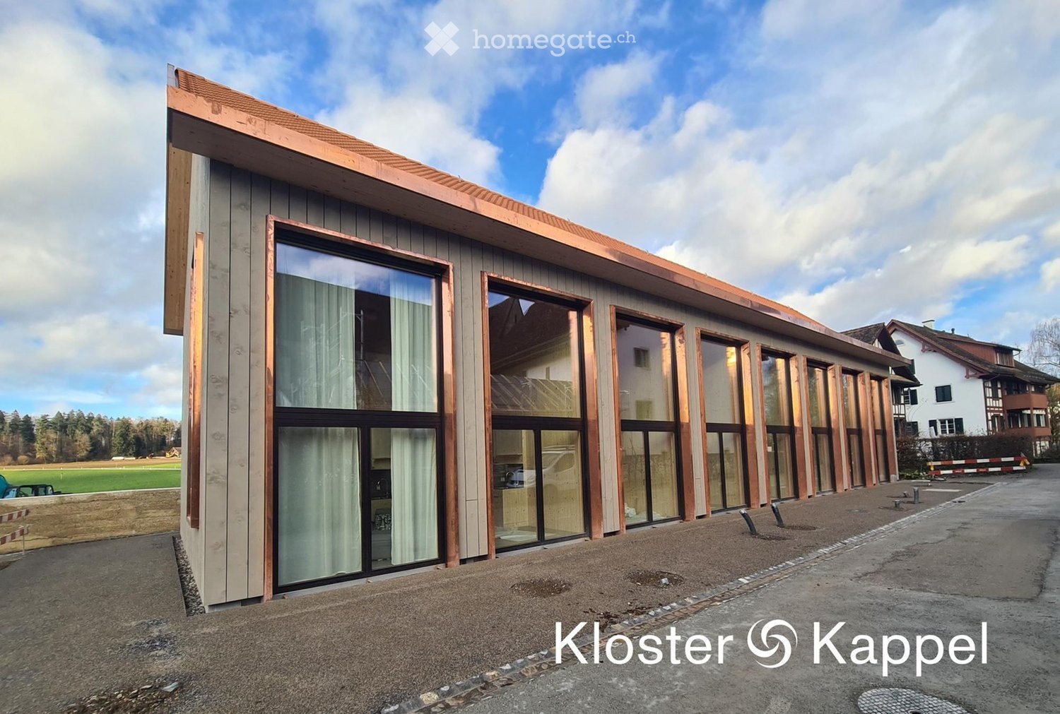 A modern building made of copper-colored metal and glass. The building has a sloped roof with a copper border, and several large windows with white curtains. The front of the building has a slanted surface with copper-colored metal strips. In the backgrou