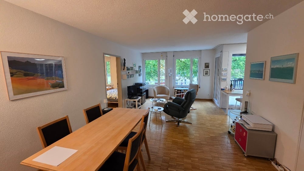 Bright, open-plan living and dining area with wooden flooring, large windows, and a balcony. Includes a dining table with chairs, a sofa, an armchair, a coffee table, a TV, and a small cabinet.