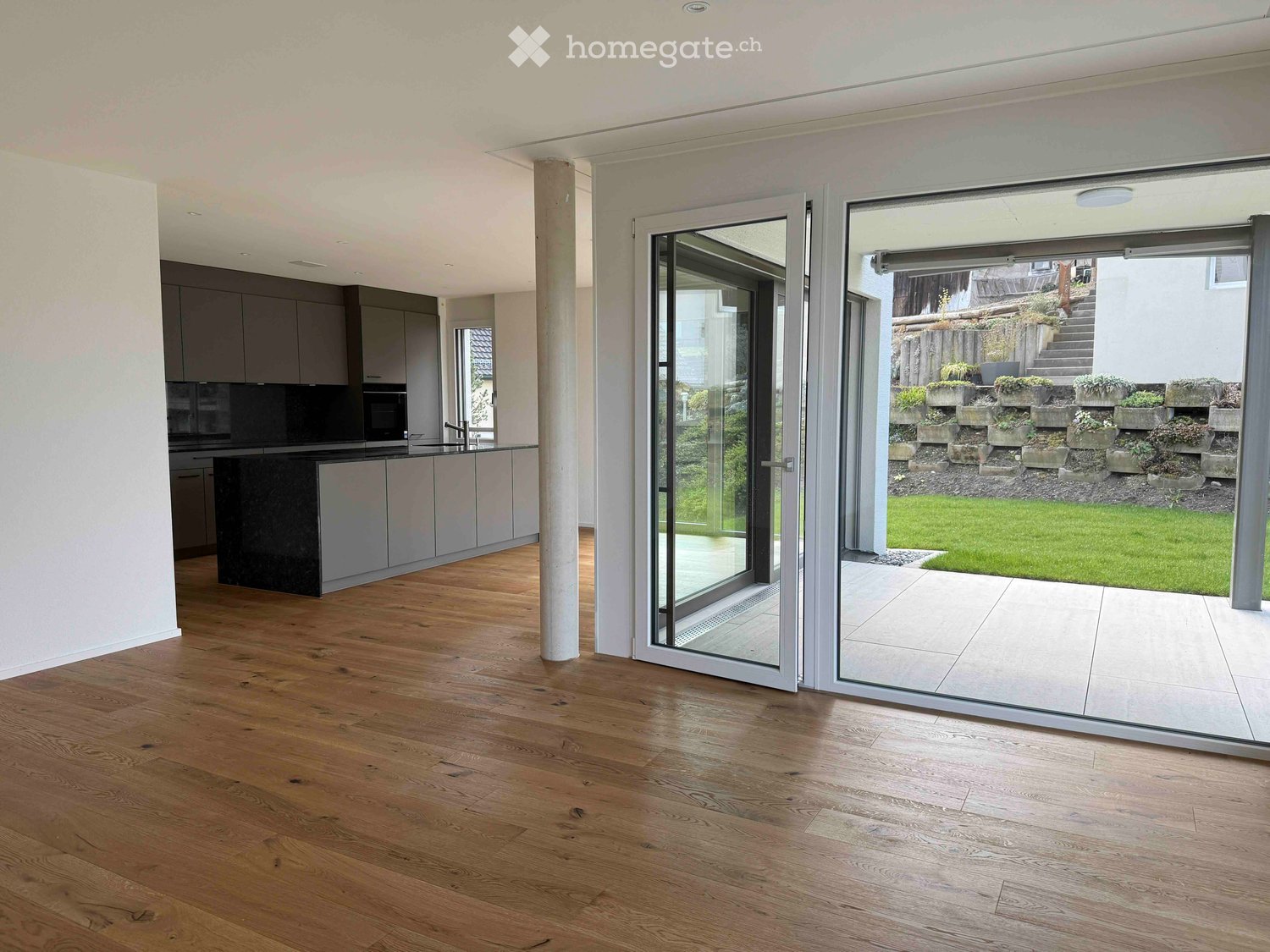 Furnished Room. Modern kitchen with light grey cabinets, dark countertop, and a large sliding glass door leading to a garden area with potted plants and a lawn.