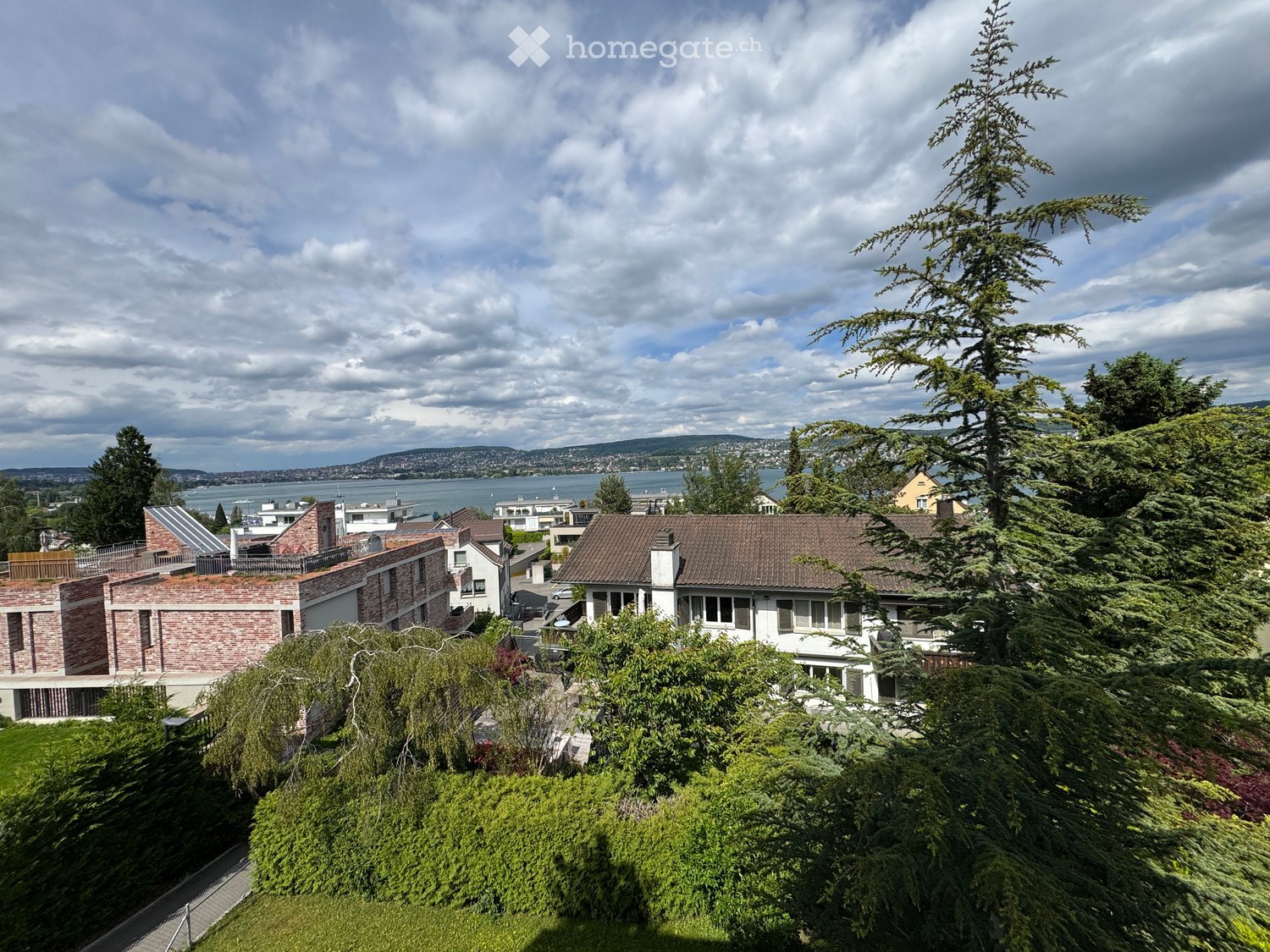 A multi-story house with a brown tiled roof, surrounded by lush greenery and trees, overlooking a body of water and distant hills.