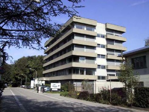 A multi-story residential building with multiple windows, two cars parked in front, two people standing in front of a sign, street lights on the side of the road, and several trees in the background.