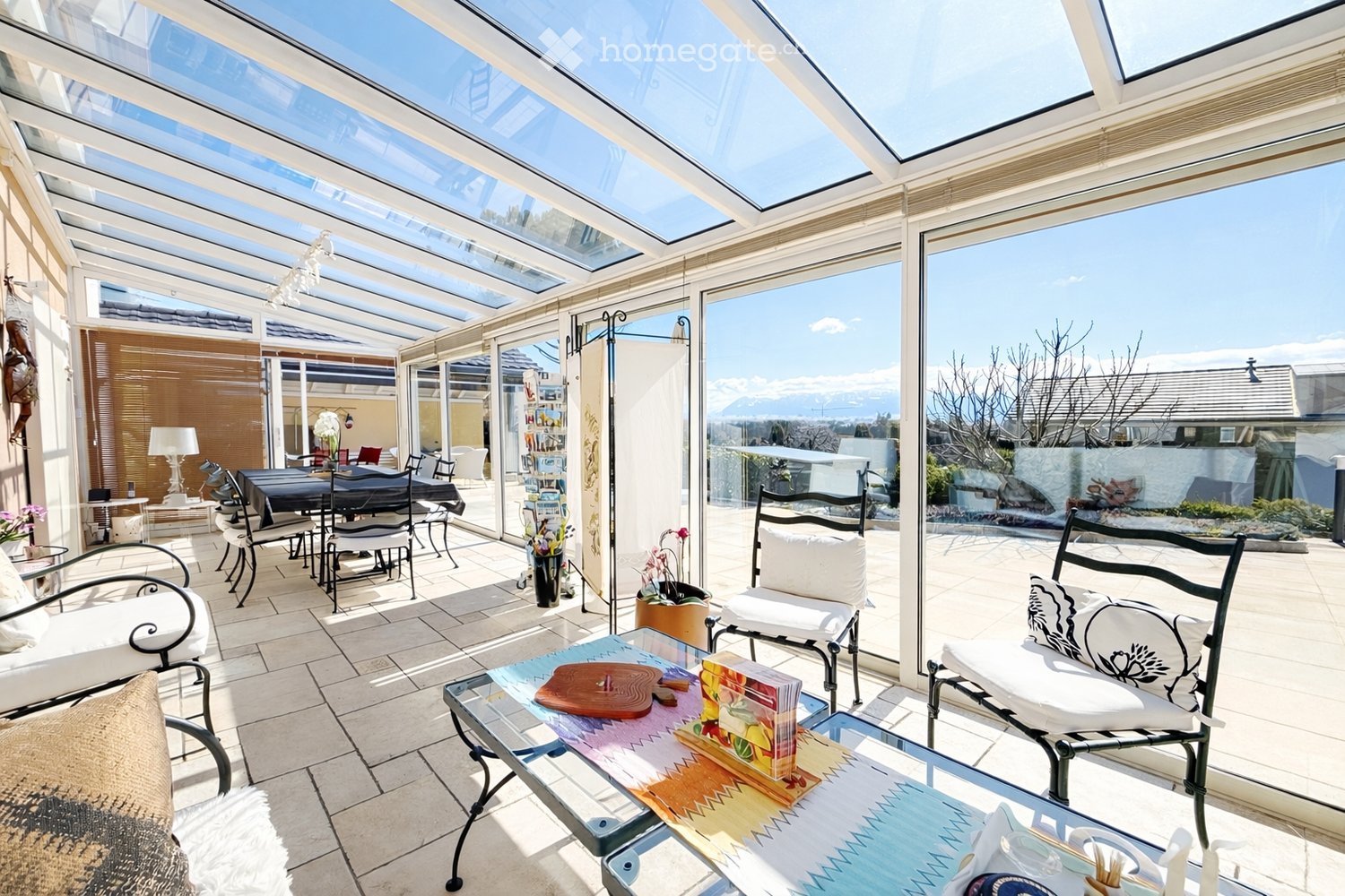 Bright, enclosed terrace with a glass roof and large windows, offering a scenic mountain view. Features a sofa, dining table, chairs, and flower pots.