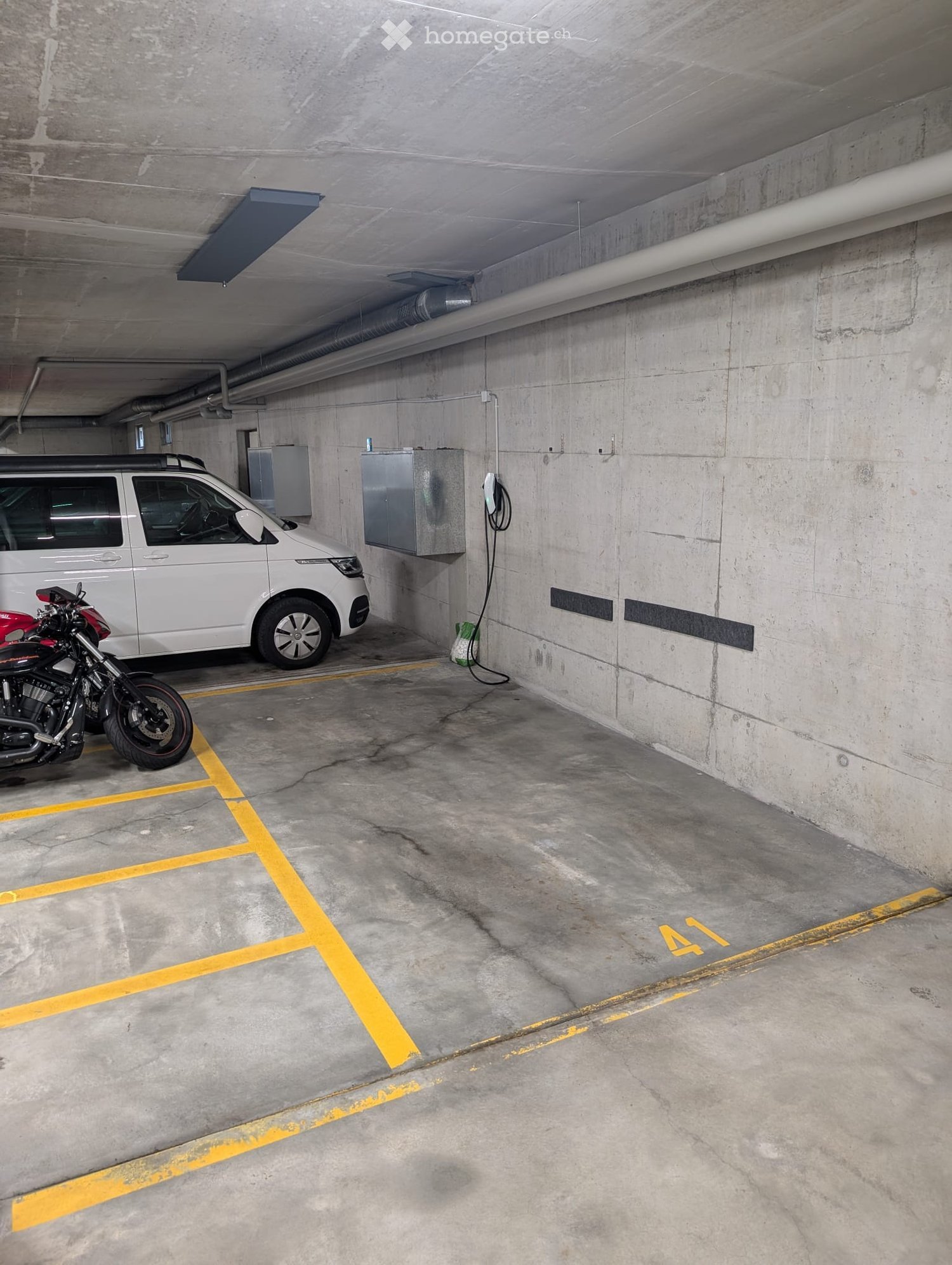 Underground parking area with a white van and a red motorcycle parked inside, marked parking spaces, and concrete walls.