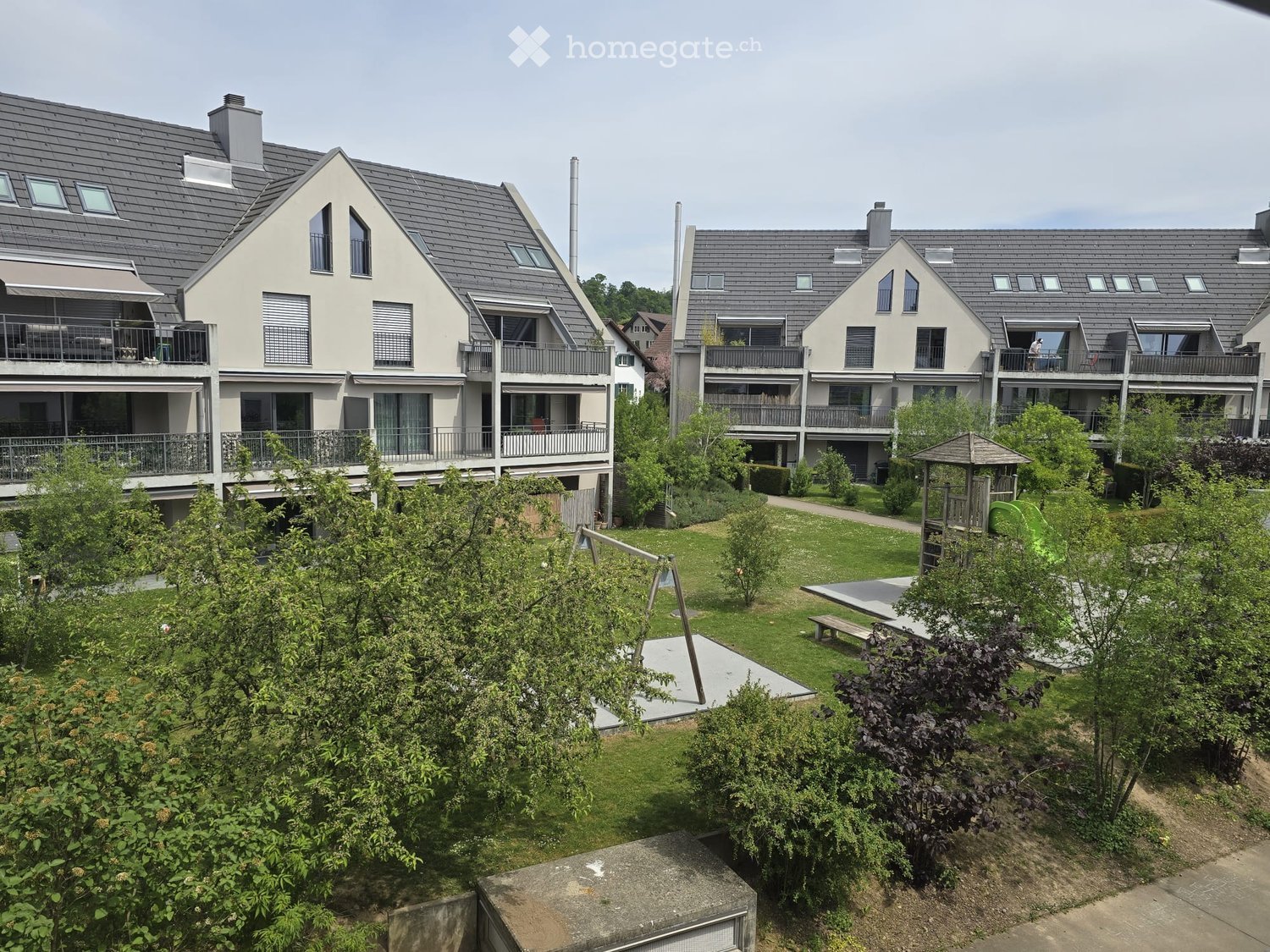 Residential complex with modern two-story houses, balconies, and a well-maintained garden area featuring a playground, benches, and trees.