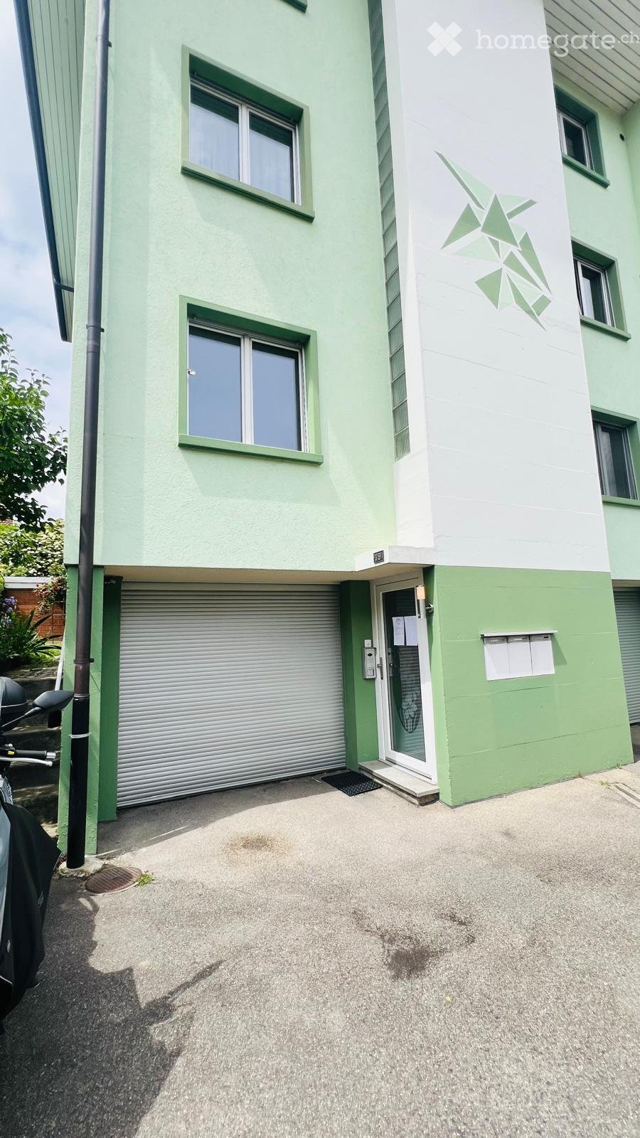 Building. Two-story building with light green and white exterior, multiple windows, garage entrance, and a small entrance door.