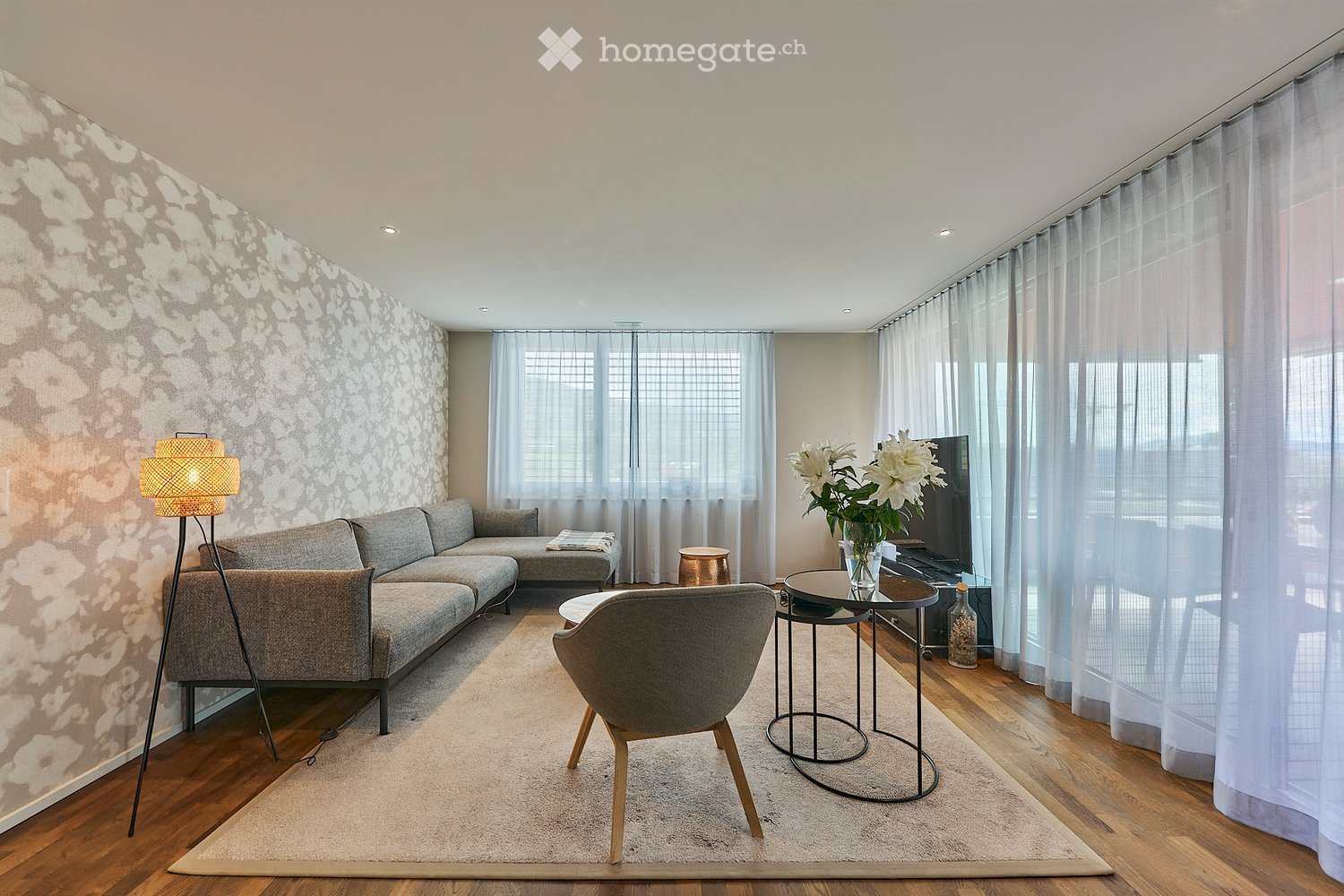 Modern living room with a grey sectional sofa, armchair, coffee table, floor lamp, and decorative vase on a side table. Large windows with sheer curtains and a patterned wall accent the space.