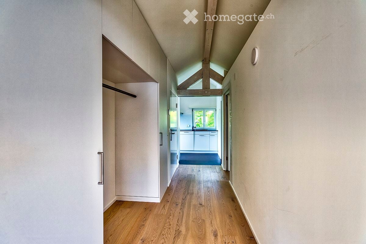 A modern hallway with wooden flooring, white walls, and built-in storage on one side. The hallway leads to a kitchen area visible at the end.