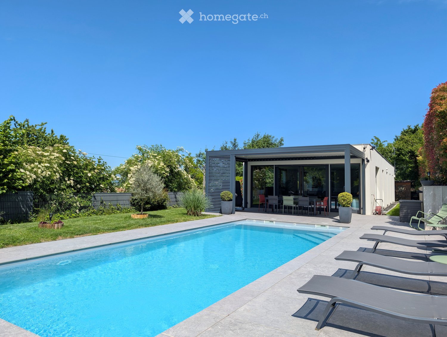 Modern outdoor area with a swimming pool, lounge chairs, and a covered patio area with dining furniture. The space is surrounded by greenery and trees.