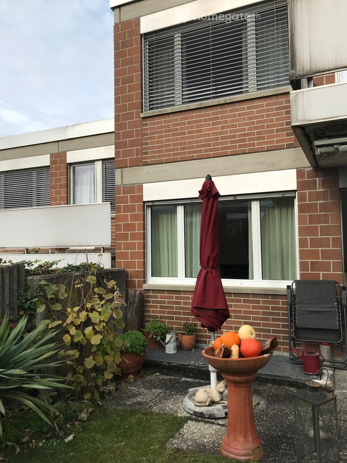 Balcony area with brick building in the background, featuring a birdbath, a small statue, a bowl with fruits, potted plants, an umbrella, and a chair.