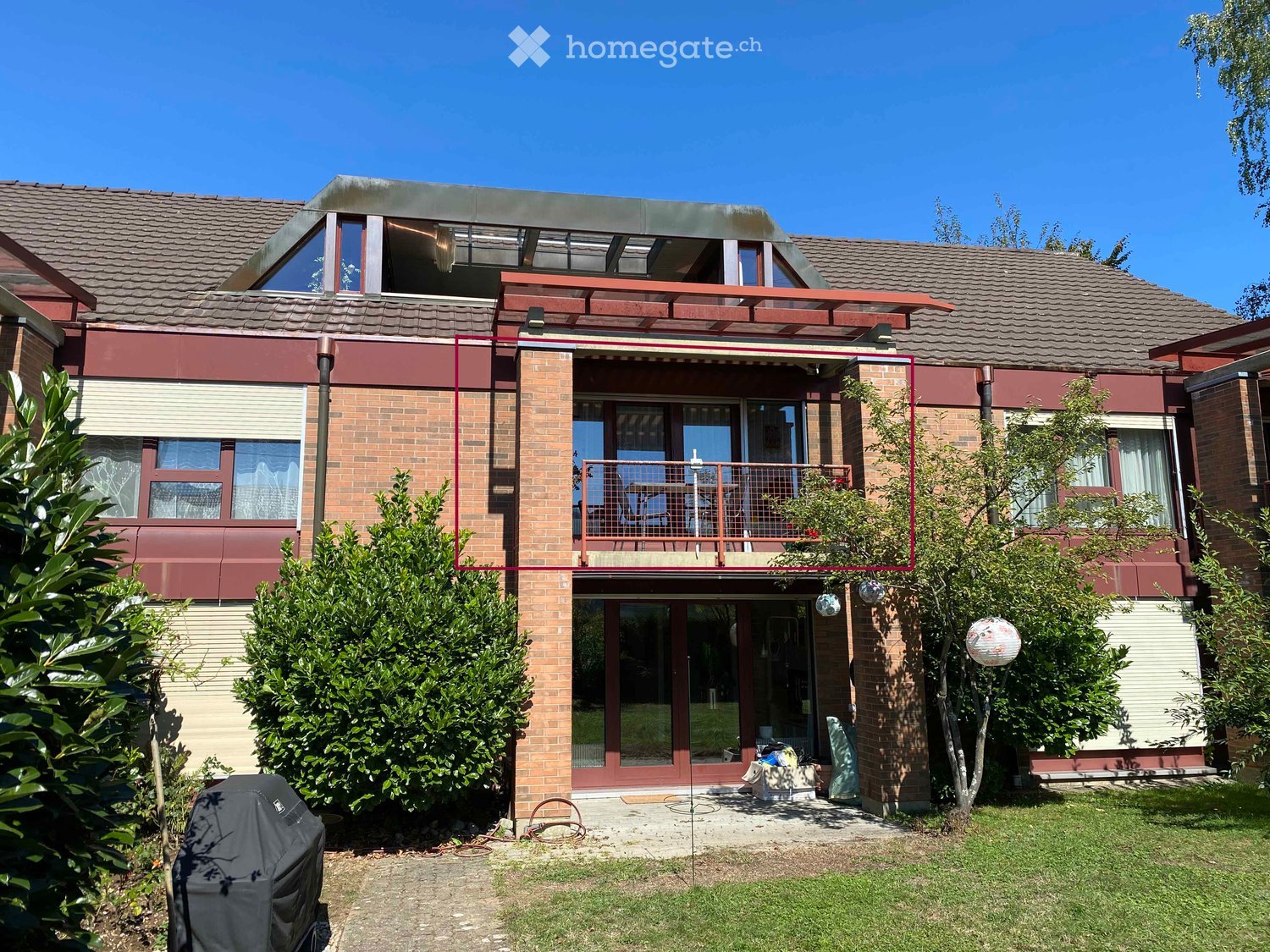 Two-story brick house with a modern design, featuring a large balcony with a railing, multiple windows, and a well-maintained garden with shrubs and a covered grill.
