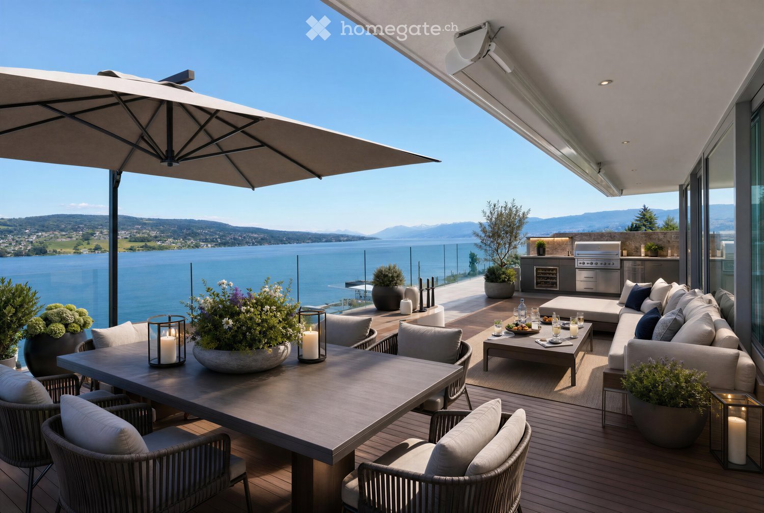 A spacious outdoor terrace with a panoramic view of a lake and mountains, featuring a large umbrella, a dining table with chairs, a sofa, a coffee table, and decorative candles and flower pots.