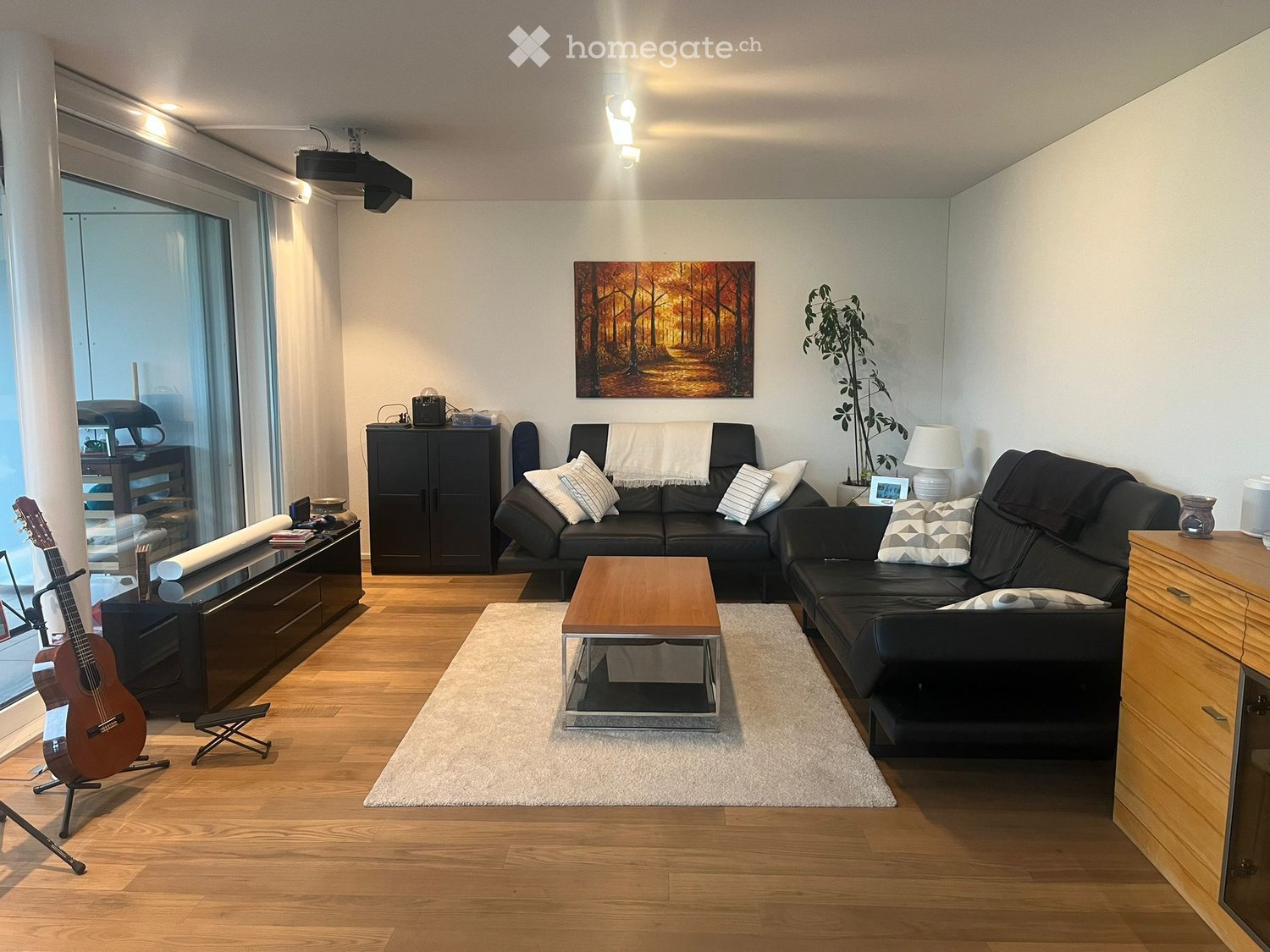 Modern living room with hardwood floors, large windows, black leather sofa, coffee table, guitar, and decorative items.