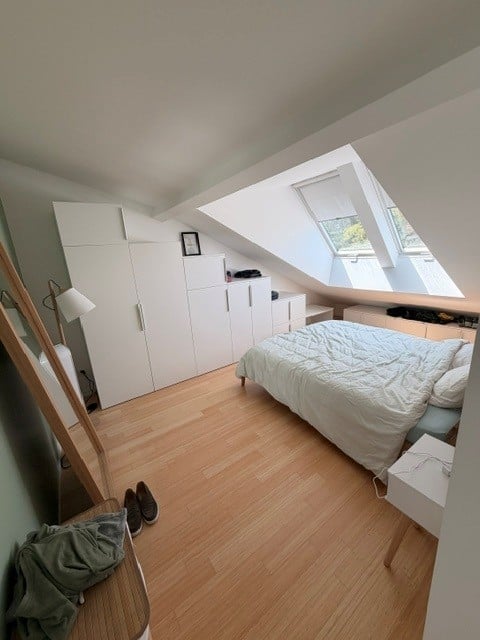 Cozy attic bedroom with wooden flooring, white wardrobe, bed with white bedding, nightstand, and a skylight window.