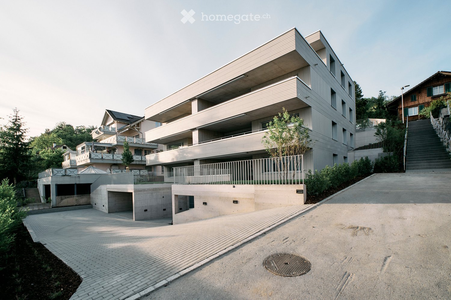 Modern multi-story residential building with clean lines, balconies, and a raised ground floor. The structure features a combination of concrete and light-colored materials, with a spacious driveway and surrounding greenery.