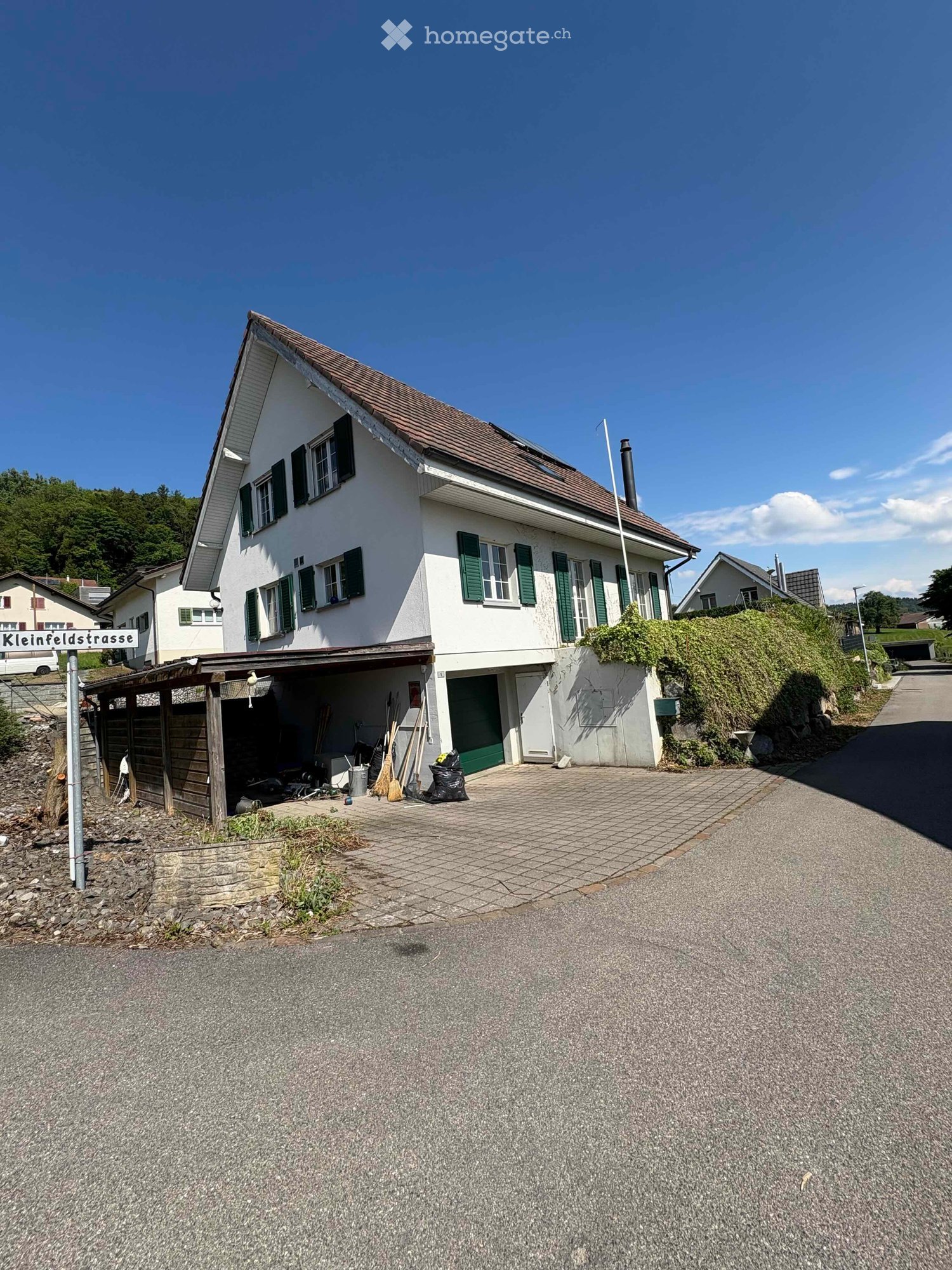Two-story white house with green shutters, brown roof, and attached garage. The house is situated on a raised ground floor with a paved driveway and a small garden area.