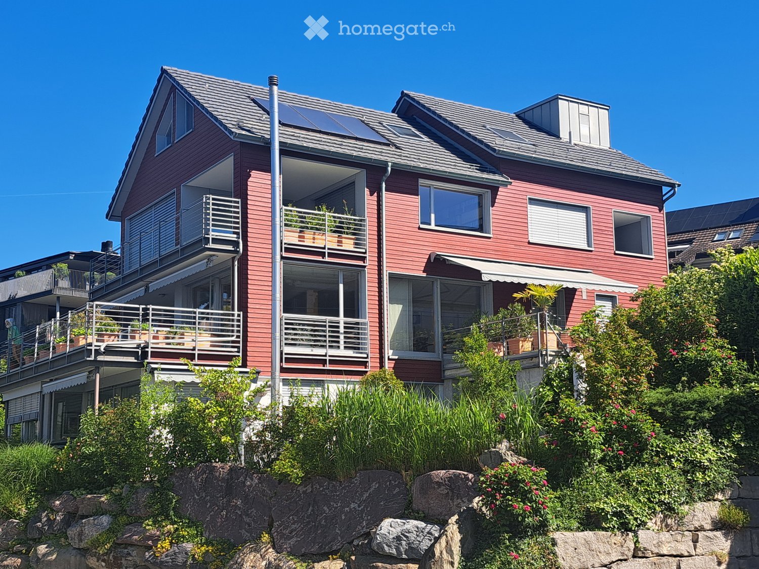 Two-story red house with multiple balconies, large windows, and solar panels on the roof. The house is surrounded by greenery and has a stone retaining wall in the foreground.